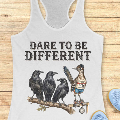 A white racerback tank top laid flat on a light wood background, featuring a graphic print of three black crows and a roadrunner wearing a race bib with the phrase 'DARE TO BE DIFFERENT'.