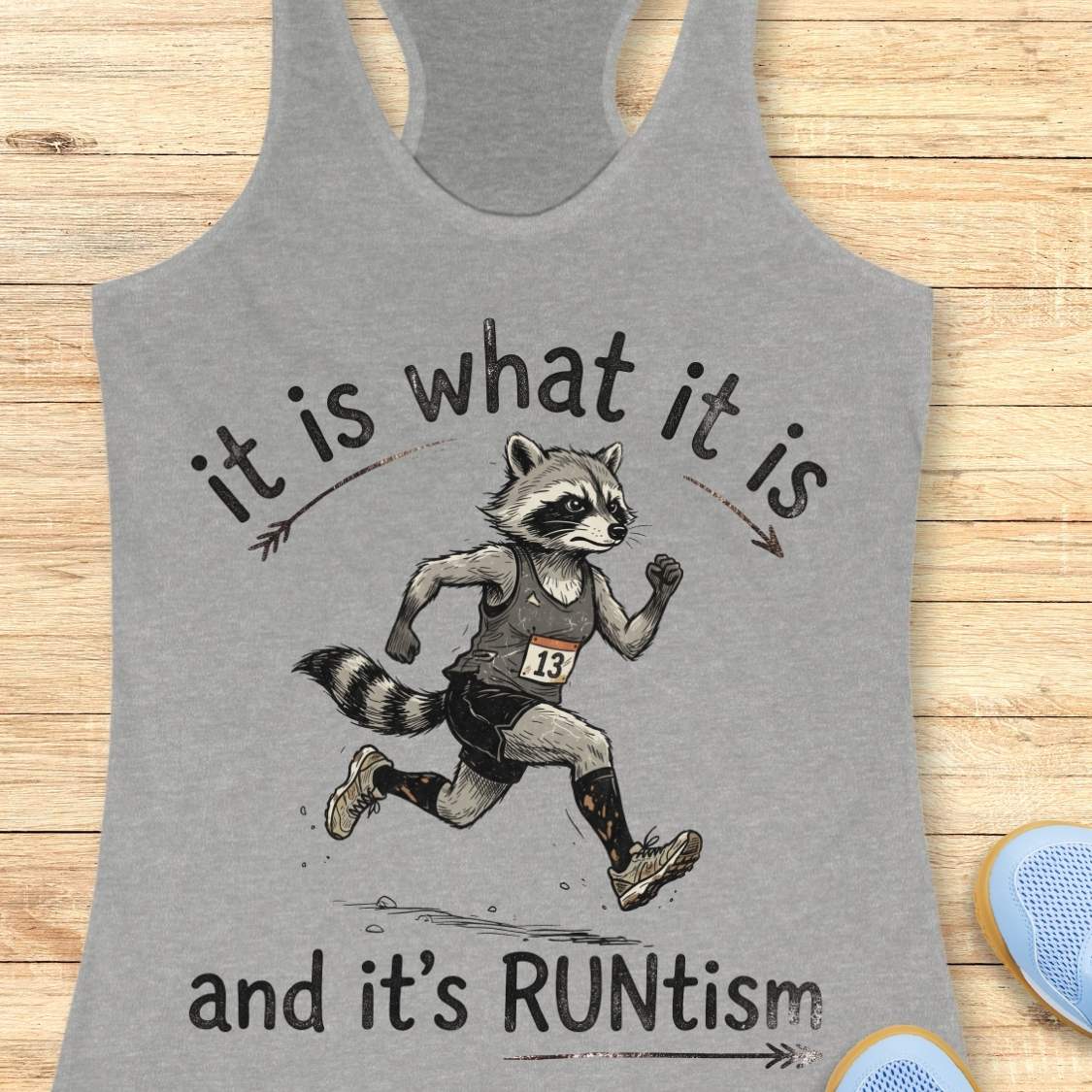A heather gray racerback tank top laid flat on a light wood surface, featuring a printed graphic of a raccoon runner with the text 'it is what it is and it's RUNtism'.