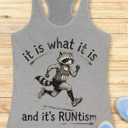 A heather gray racerback tank top laid flat on a light wood surface, featuring a printed graphic of a raccoon runner with the text 'it is what it is and it's RUNtism'.
