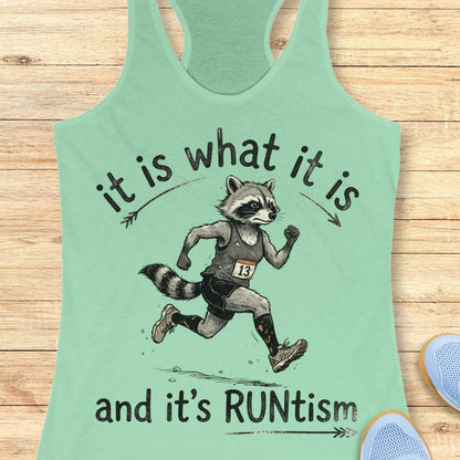 A mint green racerback tank top laid flat on a light wood background, featuring a printed graphic of a raccoon running with the text 'it is what it is and it's RUNtism'.