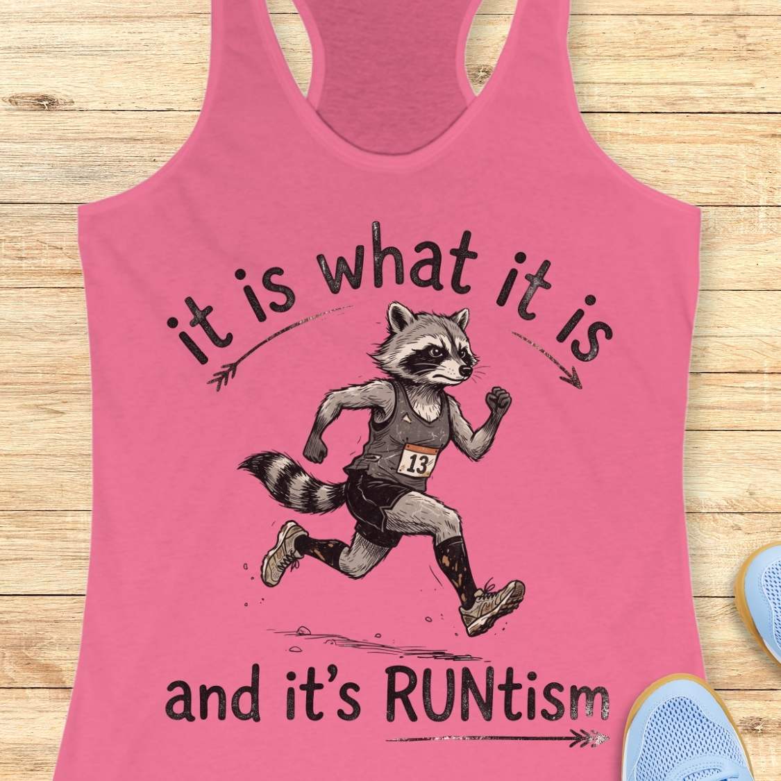 A pink racerback tank top laid flat on a light wood background, featuring a printed graphic of a raccoon running with the text 'it is what it is and it's RUNtism'.
