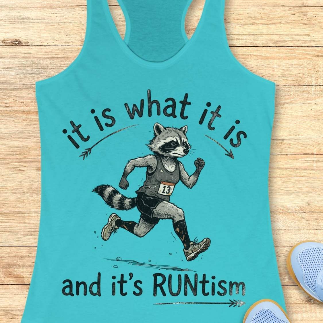 A blue racerback tank top laid flat on a light wood background, featuring a printed graphic of a raccoon running with the text 'it is what it is and it's RUNtism'.