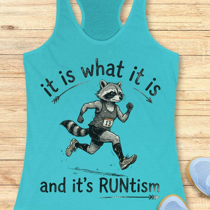 A blue racerback tank top laid flat on a light wood background, featuring a printed graphic of a raccoon running with the text 'it is what it is and it's RUNtism'.