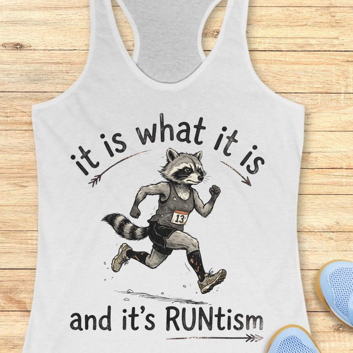 A white racerback tank top laid flat on a light wood background, featuring a printed illustration of a raccoon running with the text 'it is what it is and it's RUNtism'. Part of a blue shoe is visible at the bottom right as a styling prop.