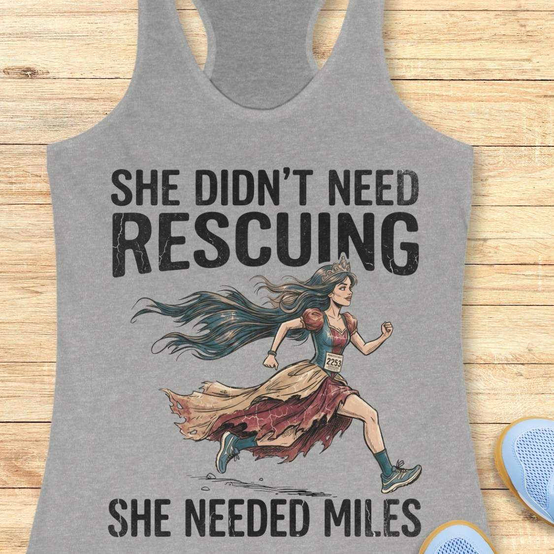 A grey racerback tank top laid flat on a light wood background, featuring a graphic of a marathon runner with the text 'SHE DIDN'T NEED RESCuing SHE NEEDED MILES'. Partial blue shoes are visible at the bottom right.