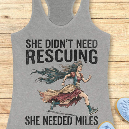 A grey racerback tank top laid flat on a light wood background, featuring a graphic of a marathon runner with the text 'SHE DIDN'T NEED RESCuing SHE NEEDED MILES'. Partial blue shoes are visible at the bottom right.