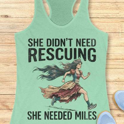 A mint racerback tank top laid flat on a light wood background, featuring a graphic of a marathon runner with the text 'SHE DIDN'T NEED RESCuing SHE NEEDED MILES'. Partial blue shoes are visible at the bottom right.