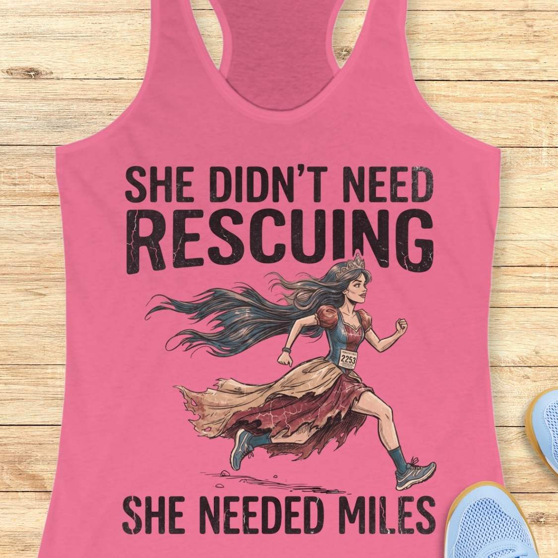 A pink racerback tank top laid flat on a light wood background, featuring a graphic of a marathon runner with the text 'SHE DIDN'T NEED RESCuing SHE NEEDED MILES'. Partial blue shoes are visible at the bottom right.