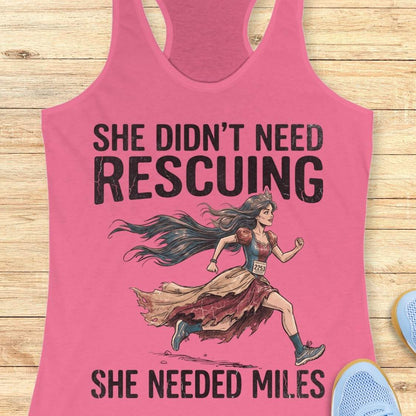 A pink racerback tank top laid flat on a light wood background, featuring a graphic of a marathon runner with the text 'SHE DIDN'T NEED RESCuing SHE NEEDED MILES'. Partial blue shoes are visible at the bottom right.