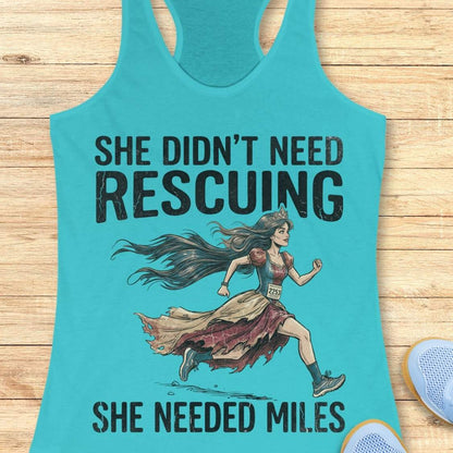 A turquoise racerback tank top laid flat on a light wood surface, featuring a distressed black graphic and text that reads 'SHE DIDN'T NEED RESCuing SHE NEEDED MILES' with an illustrated female runner in a torn dress. A partial blue shoe is visible at the lower right edge.