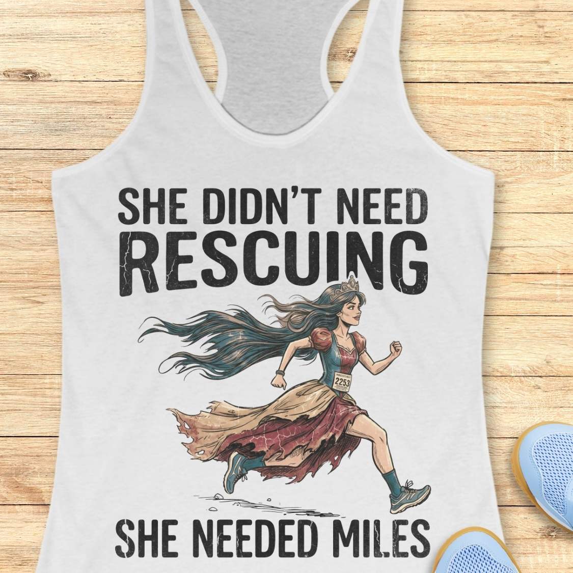A white racerback tank top laid flat on a light wood background, featuring a graphic of a marathon runner with the text 'SHE DIDN'T NEED RESCuing SHE NEEDED MILES'. Partial blue shoes are visible at the bottom right.