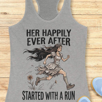 A heather gray racerback tank top laid flat on a light wood background, featuring a printed graphic of a woman running with the text 'HER HAPPILY EVER AFTER STARTED WITH A RUN'.
