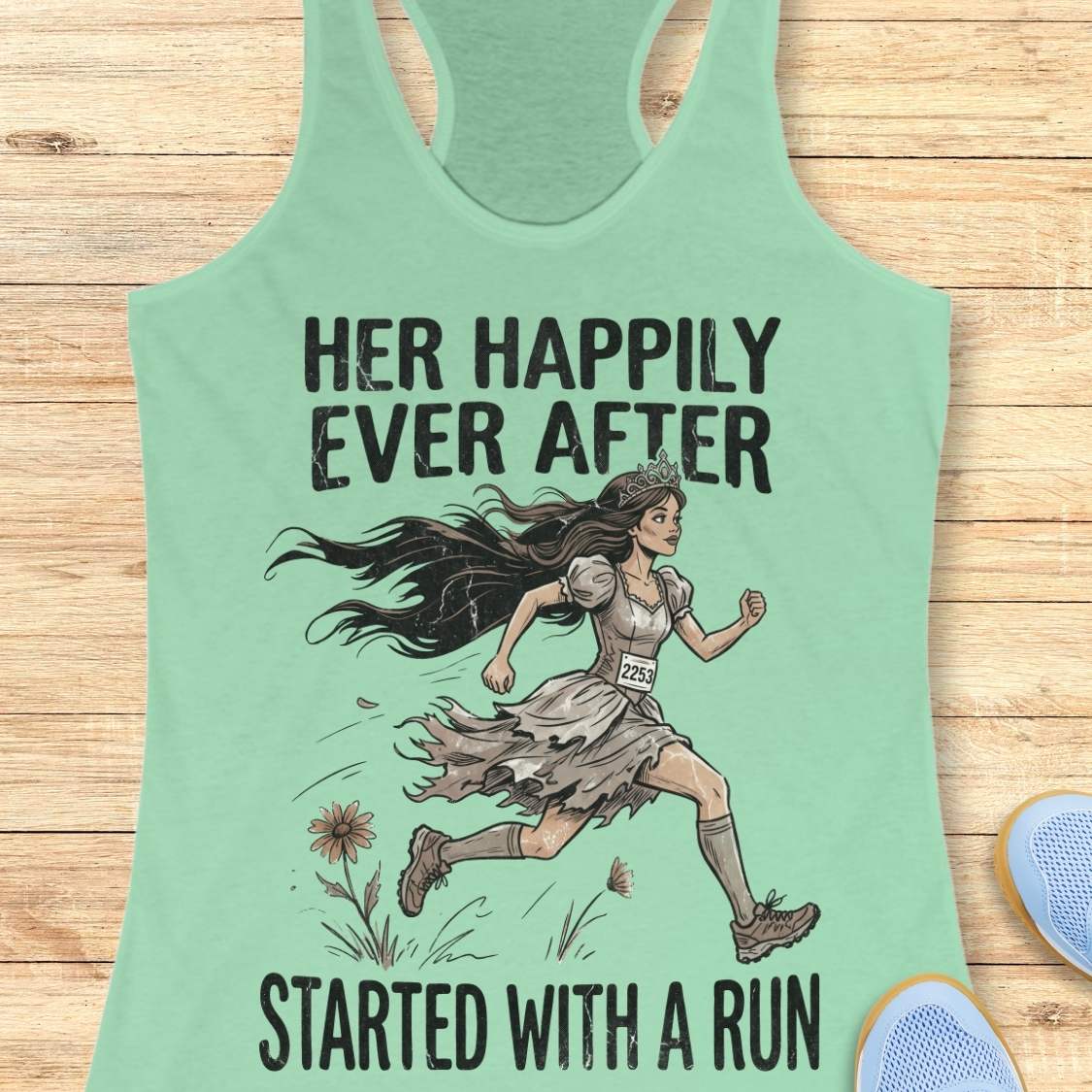 A mint racerback tank top laid flat on a light wood background, featuring a printed graphic of a woman running with the text 'HER HAPPILY EVER AFTER STARTED WITH A RUN'.