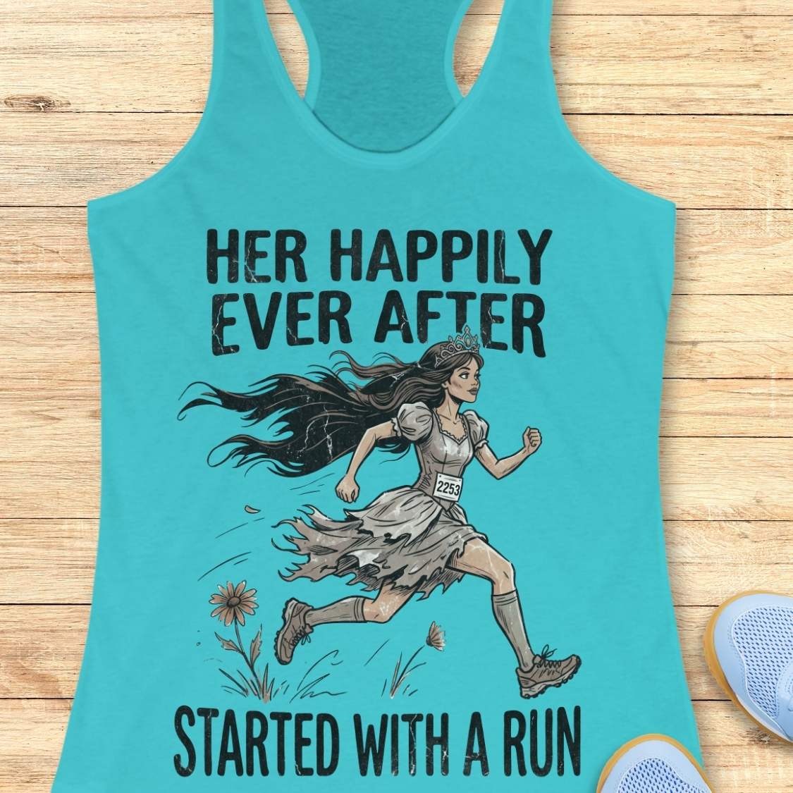 A turquoise racerback tank top laid flat on a light wood surface, featuring a printed illustration of a woman running with the text 'HER HAPPILY EVER AFTER STARTED WITH A RUN.' A pair of blue shoes is partially visible at the lower right edge.