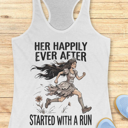 A white racerback tank top laid flat on a light wood background, featuring a graphic of a woman runner with the text 'HER HAPPILY EVER AFTER STARTED WITH A RUN'. Partial blue shoes are visible at the bottom right as styling props.