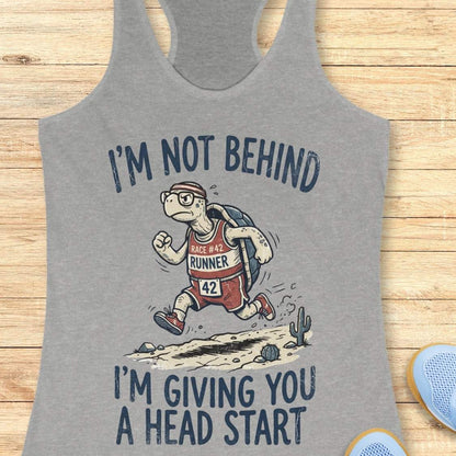 A heather gray racerback tank top laid flat on a light wood background, featuring a cartoon turtle runner graphic with the text 'I'M NOT BEHIND I'M GIVING YOU A HEAD START'.