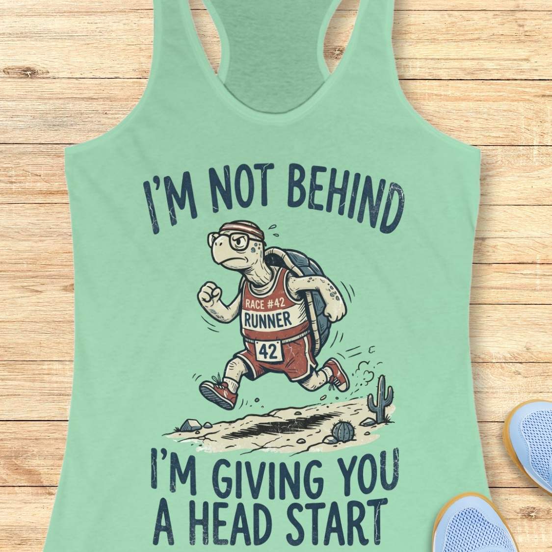 A mint green racerback tank top laid flat on a light wood background, featuring a cartoon turtle running with the text 'I'M NOT BEHIND I'M GIVING YOU A HEAD START'.