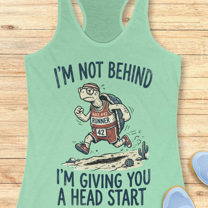 A mint green racerback tank top laid flat on a light wood background, featuring a cartoon turtle running with the text 'I'M NOT BEHIND I'M GIVING YOU A HEAD START'.