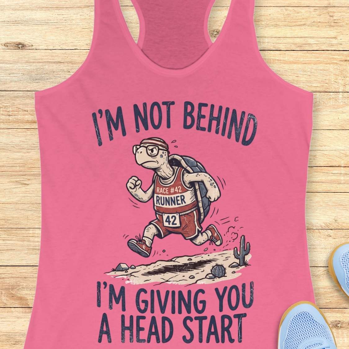 A pink racerback tank top laid flat on a light wood surface, featuring a cartoon turtle runner graphic with the text 'I'M NOT BEHIND I'M GIVING YOU A HEAD START'. Partial blue shoes are visible at the edges as styling props.