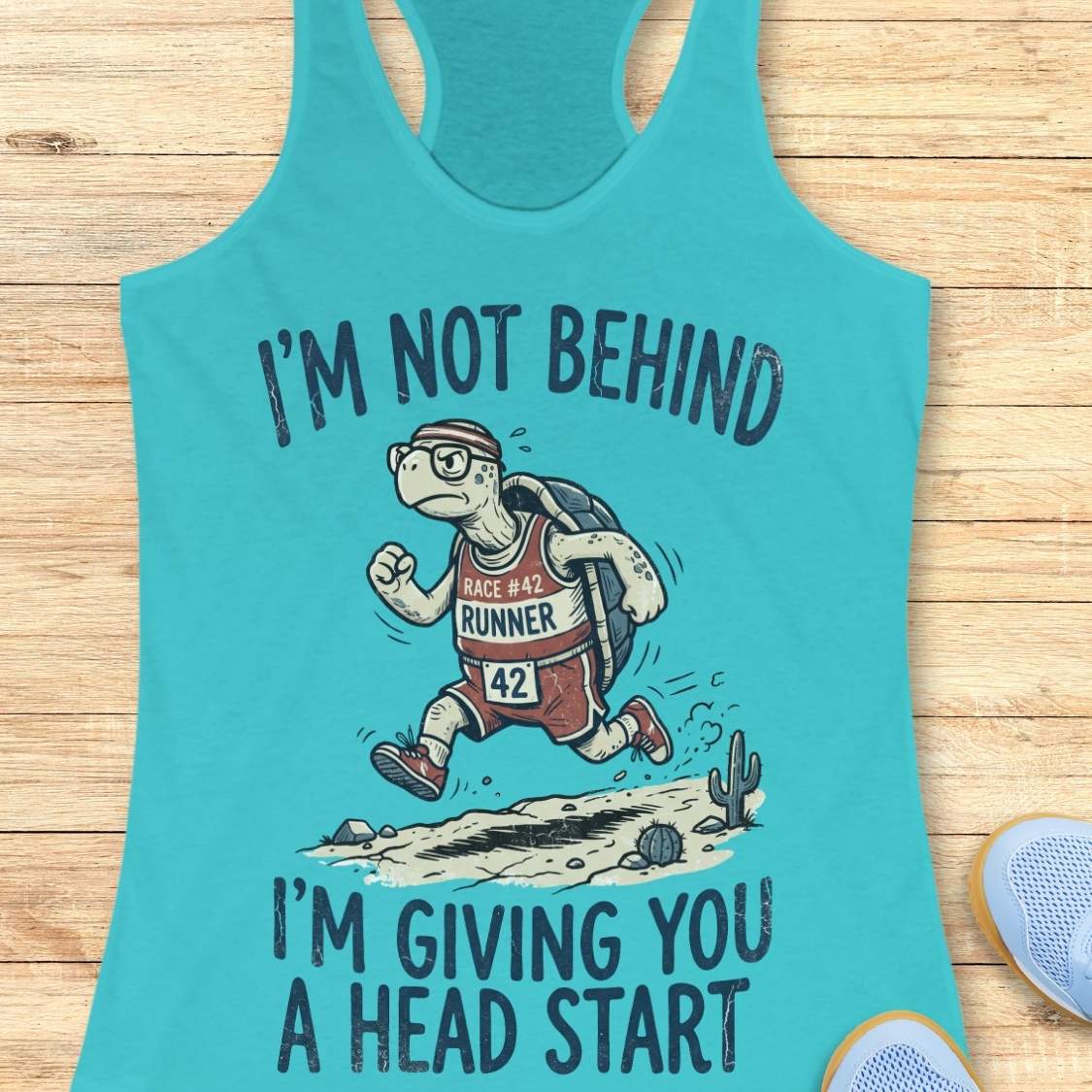 A turquoise racerback tank top laid flat on a light wood surface, featuring a cartoon turtle runner graphic with the text 'I'M NOT BEHIND I'M GIVING YOU A HEAD START'. Partial light blue shoes are visible at the bottom right as styling props.