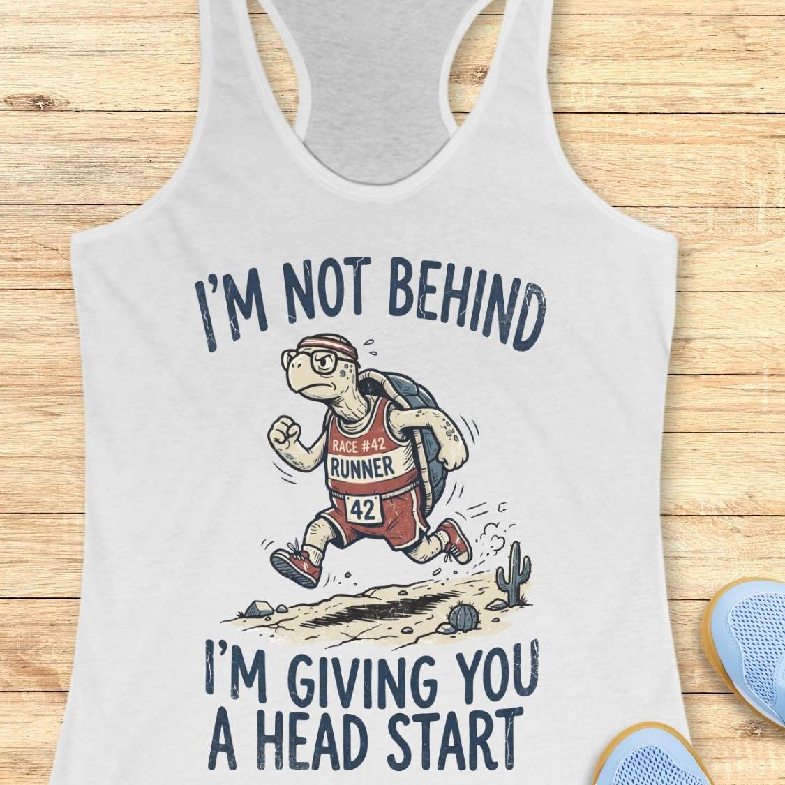 A white racerback tank top laid flat on a light wood background, featuring a graphic of a cartoon turtle running with the text 'I'M NOT BEHIND I'M GIVING YOU A HEAD START'. Partial blue shoes are visible at the bottom right edge.