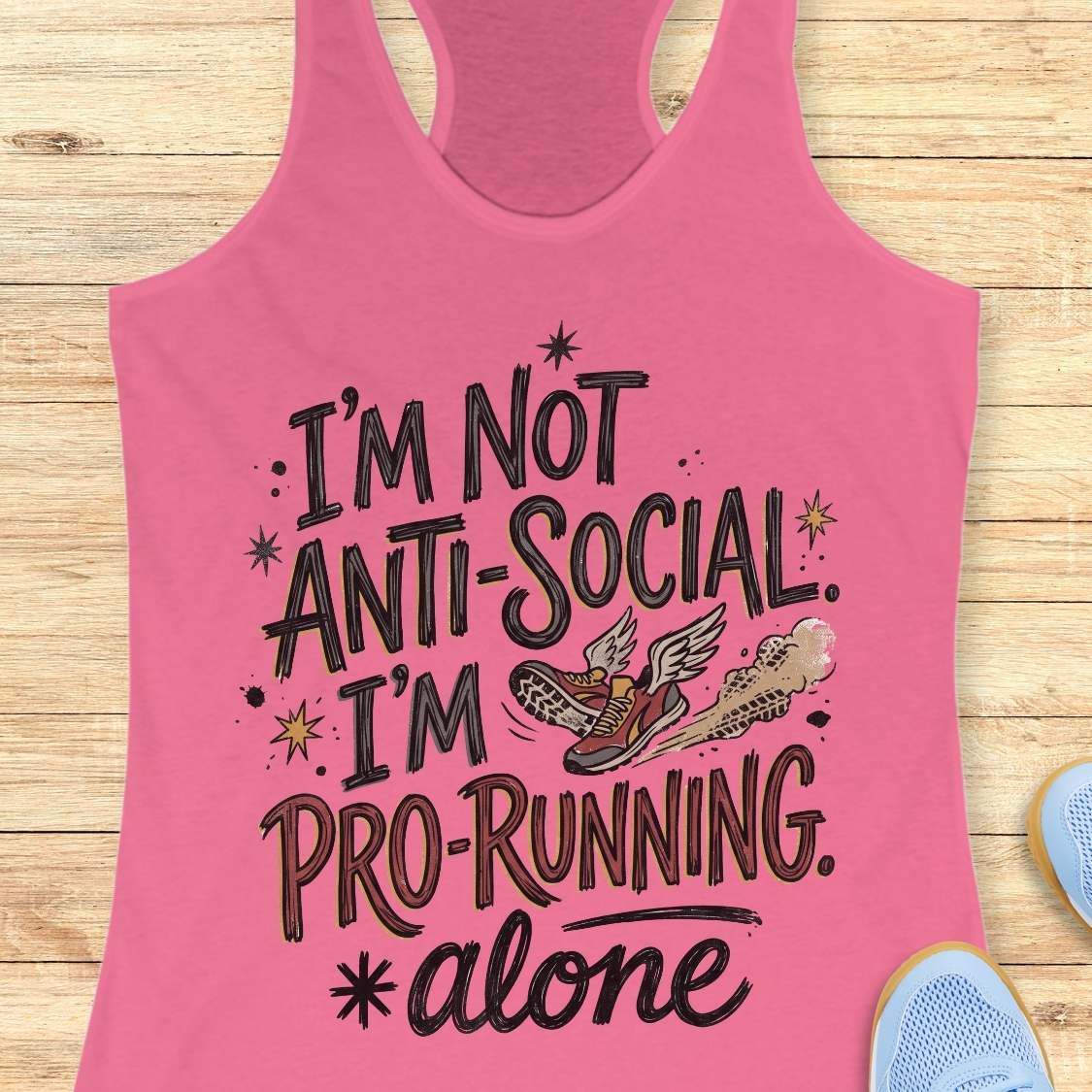 A pink racerback tank top laid flat on a light wood background, featuring the printed phrase “I’m Not Anti-Social. I’m Pro-Running.