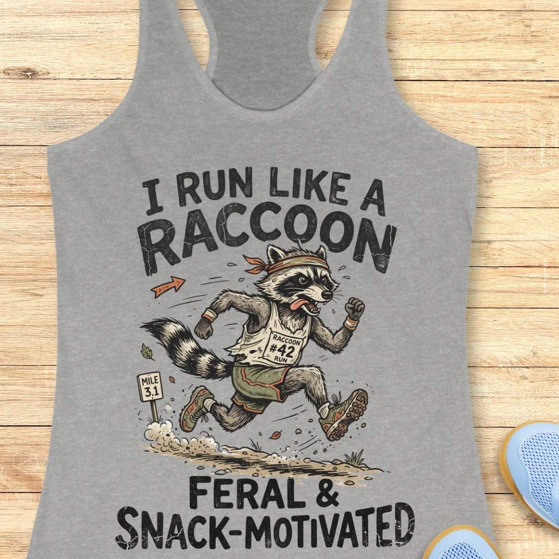A heather gray racerback tank top laid flat on a light wood background, featuring a humorous graphic of a raccoon running a race with the text 'I RUN LIKE A RACCOON' and 'FERAL & SNACK-MOTIVATED'.