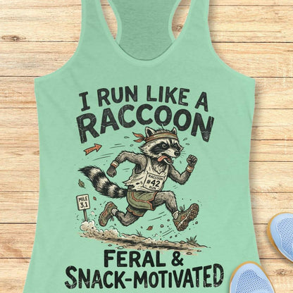 A mint green racerback tank top laid flat on a light wood background, featuring a humorous graphic of a raccoon running a race with the text 'I RUN LIKE A RACCOON FERAL & SNACK-MOTIVATED'.