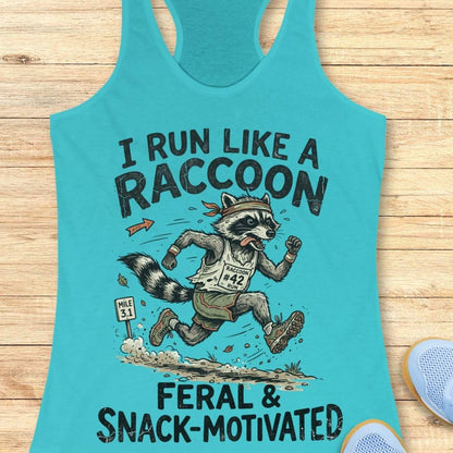 A turquoise racerback tank top laid flat on a light wood surface, featuring a humorous raccoon race graphic with the text 'I RUN LIKE A RACCOON' and 'FERAL & SNACK-MOTIVATED.' A small blue shoe is partially visible at the lower right edge.