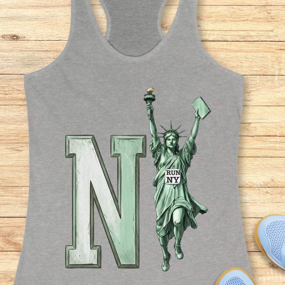 A heather gray racerback tank top laid flat on a light wood background, featuring a large mint green letter 'N' and a Statue of Liberty graphic with 'RUN NY' printed on the robe.