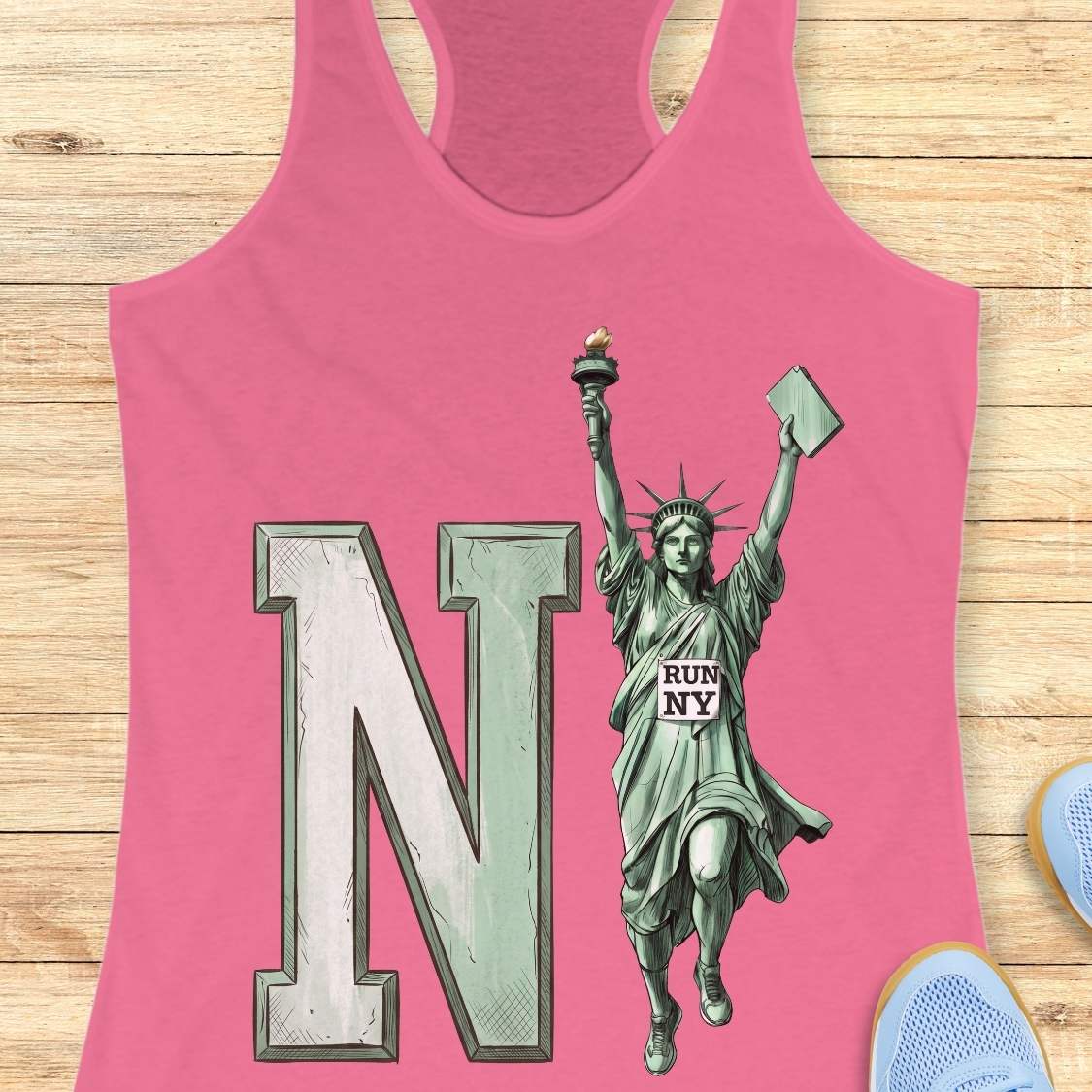 A pink racerback tank top laid flat on a light wood background, featuring a large distressed letter N and a Statue of Liberty graphic with the words 'RUN NY'.