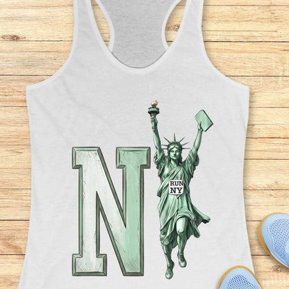 A white racerback tank top laid flat on a light wood background, featuring a large mint green letter 'N' and a Statue of Liberty graphic with 'RUN NY' printed on the statue's chest. Partial blue shoes are visible at the bottom right edge.