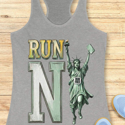 A heather gray racerback tank top laid flat on a light wood background, featuring a large graphic print with the word 'RUN', a large letter 'N', and an illustrated Statue of Liberty running with a 'RUN FREE' sign.