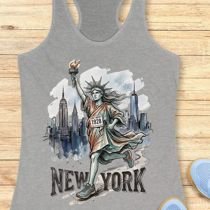 A heather gray racerback tank top featuring a graphic of the Statue of Liberty running with the New York City skyline behind her and the text 'NEW YORK' at the bottom. The tank top is displayed flat on a light wood background.