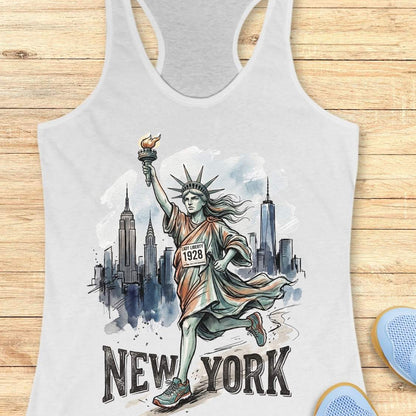 A white racerback tank top laid flat on a light wood surface, featuring a watercolor-style graphic of the Statue of Liberty running with the New York City skyline behind her and the text 'NEW YORK' below. Part of a blue shoe is visible at the bottom right.
