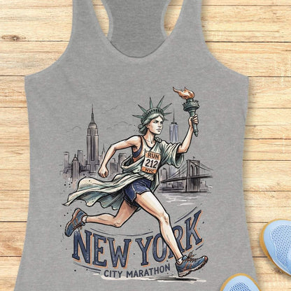 A heather gray racerback tank top featuring a detailed graphic of a woman running the New York City Marathon, holding a torch, with the Statue of Liberty and New York City skyline in the background. The tank top is displayed flat on a light brown wooden surface.
