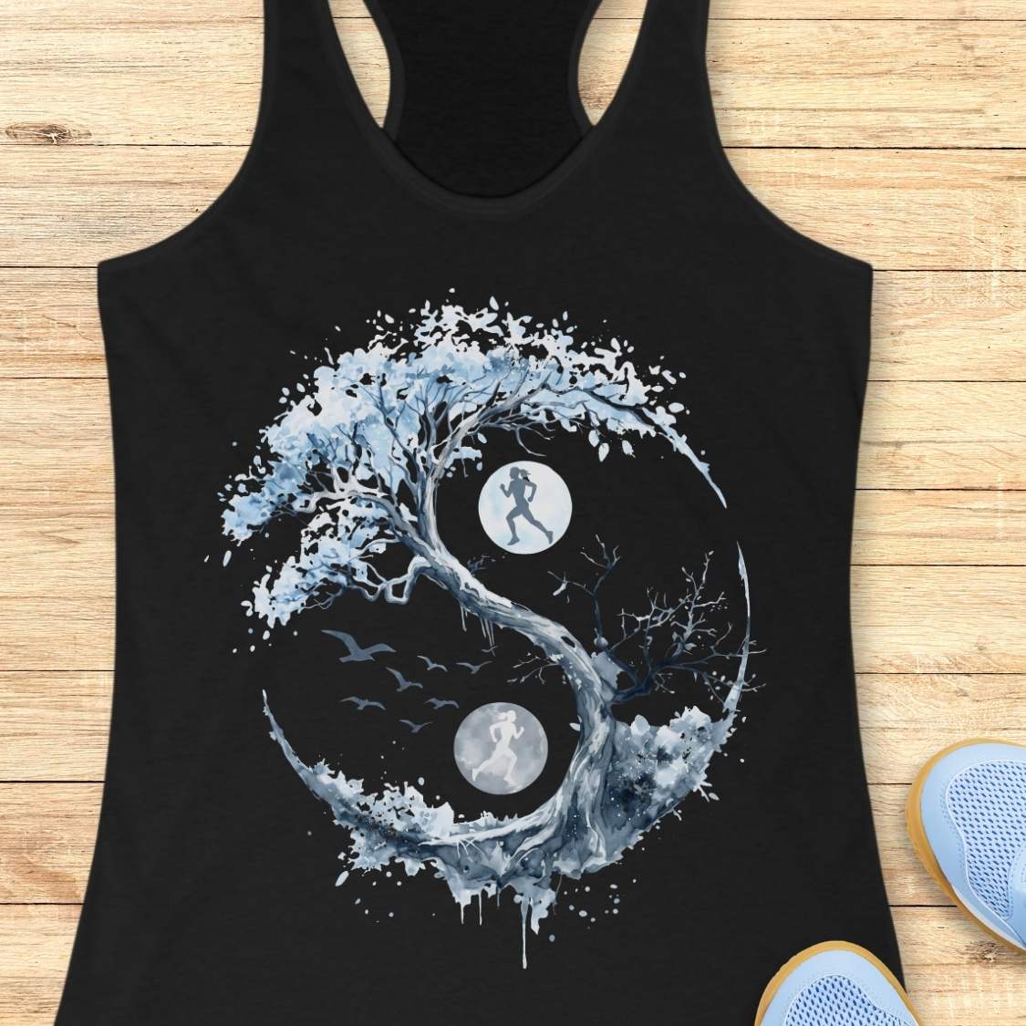 A black racerback tank top laid flat on a light wood floor, featuring a large yin-yang style graphic made from a tree, birds, and two moon phases with running silhouettes. Partial light blue sneakers are visible at the bottom right as styling props.