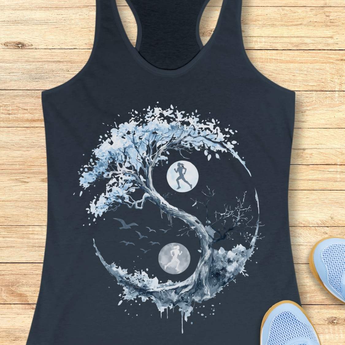 A indigo racerback tank top laid flat on a light wood floor, featuring a large yin-yang style graphic made from a tree, birds, and two moon phases with running silhouettes. Partial light blue sneakers are visible at the bottom right as styling props.