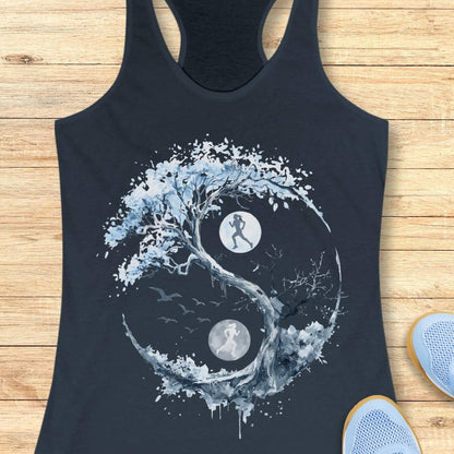 A indigo racerback tank top laid flat on a light wood floor, featuring a large yin-yang style graphic made from a tree, birds, and two moon phases with running silhouettes. Partial light blue sneakers are visible at the bottom right as styling props.