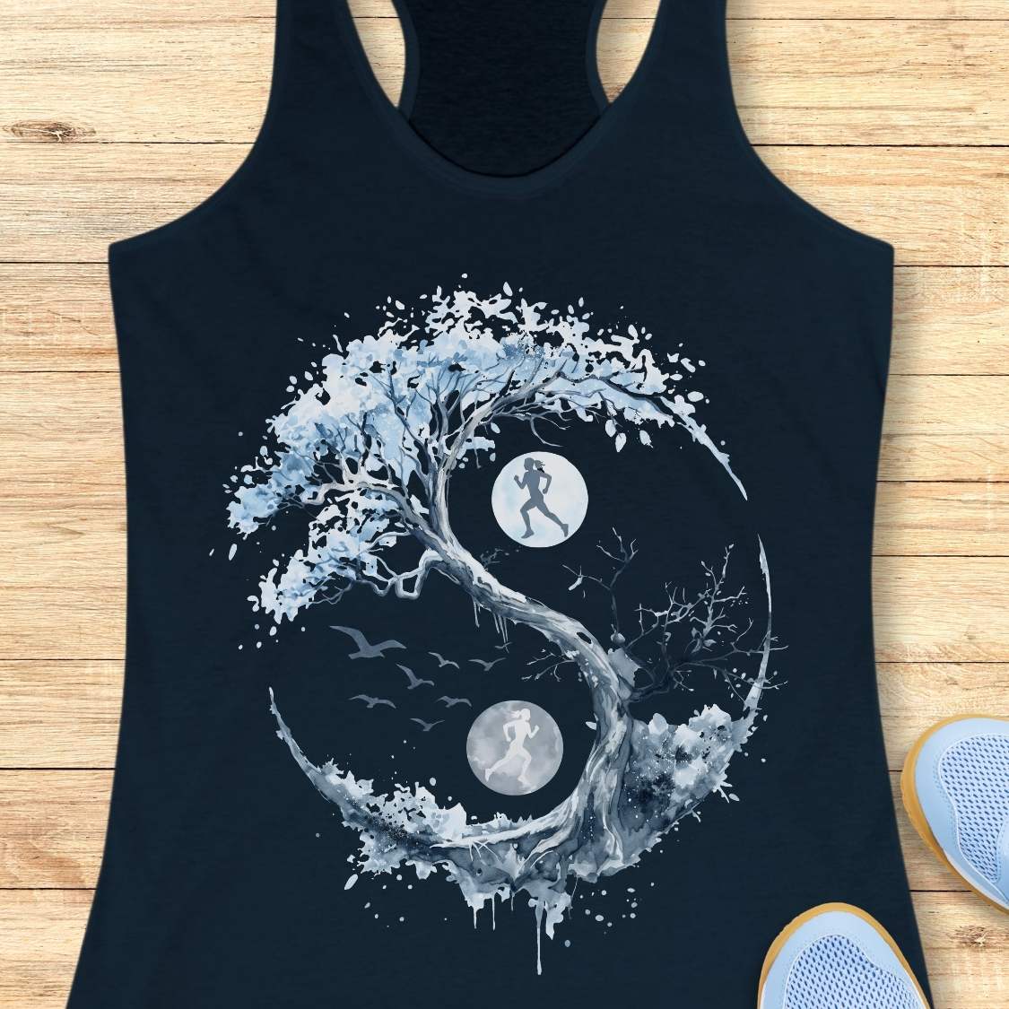 A navy racerback tank top laid flat on a light wood surface, featuring a large white and light blue yin-yang style graphic with tree branches, birds, and two circular moon/sun elements. Part of a light blue shoe is visible at the lower right edge.