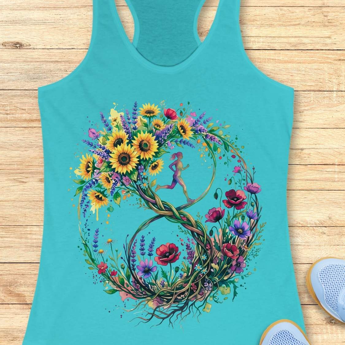 A turquoise racerback tank top laid flat on a light wood surface, featuring a large colorful floral and vine graphic with sunflowers, red poppies, purple wildflowers, and a small running fairy motif. Partial light blue sneakers are visible at the bottom right as styling props.