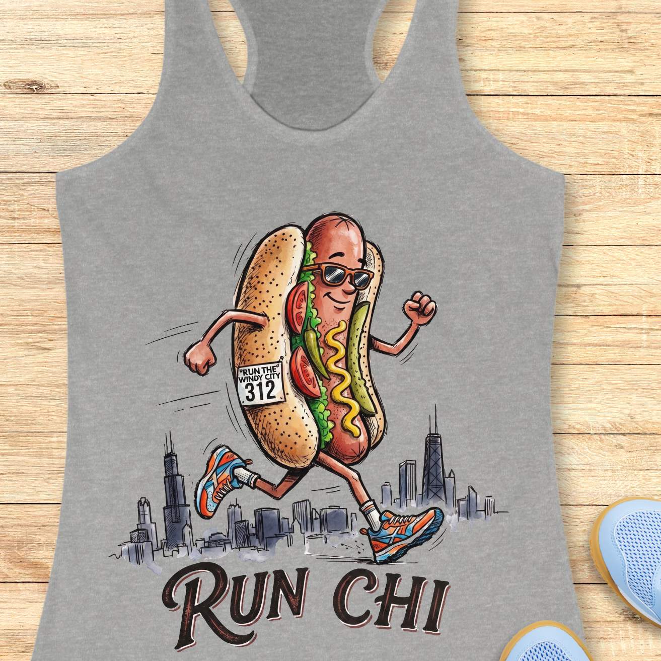heather grey women's racerback tank top featuring a playful illustration of a running hot dog wearing sunglasses and colorful running shoes. The hot dog has a race bib that reads “RUN THE WINDY CITY 312” and is jogging in front of a hand-drawn Chicago skyline. The design includes classic Chicago-style hot dog toppings such as tomatoes, pickles, mustard, and relish. Large text below the illustration reads: “RUN CHI.” The artwork combines running humor with Chicago pride in a fun, cartoon-style design.
