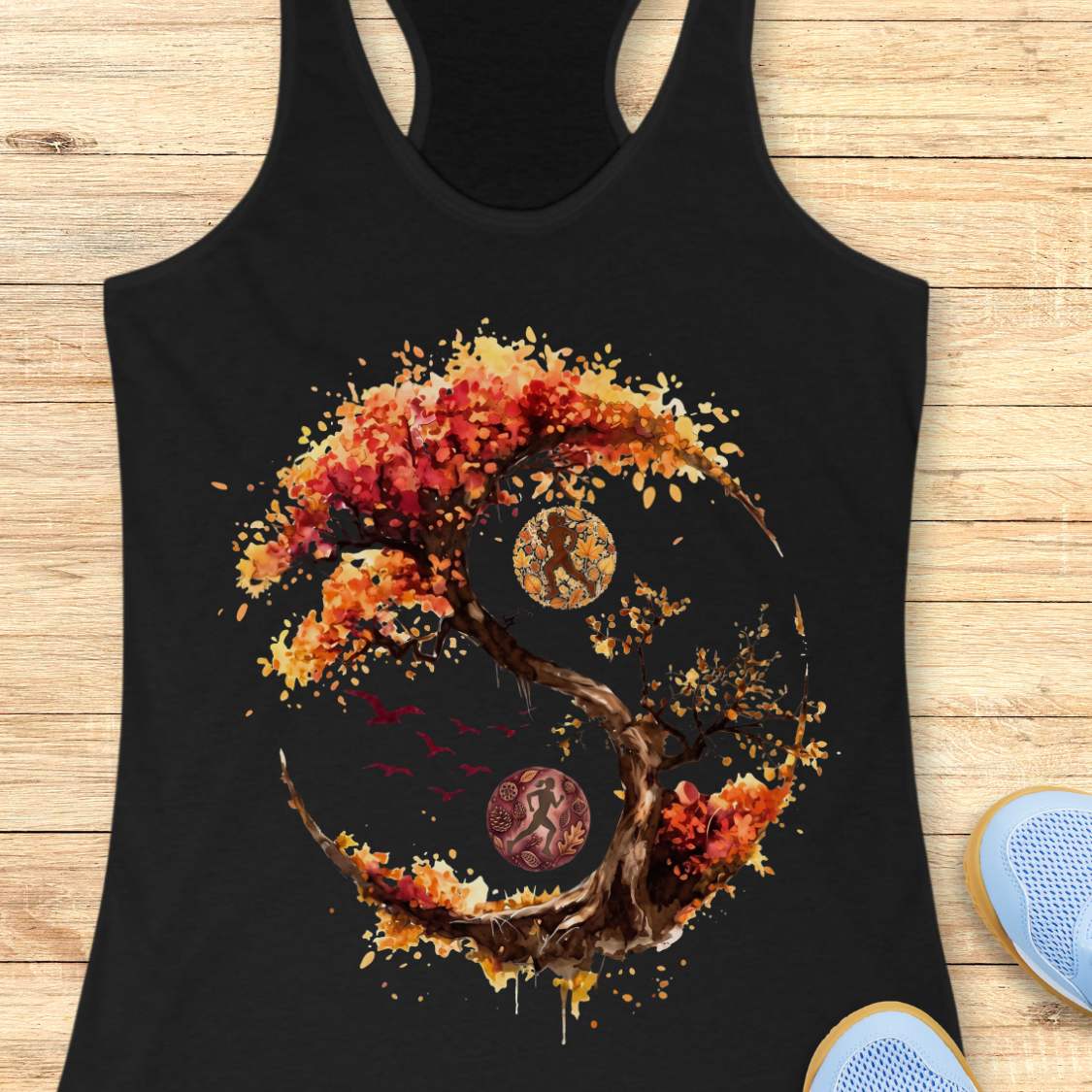 A black racerback tank top laid flat on a light wood plank background, featuring a large graphic print of a stylized tree in autumn colors forming a circular yin-yang shape with a running figure, birds, and circular motifs. Part of a blue shoe is visible at the bottom right.