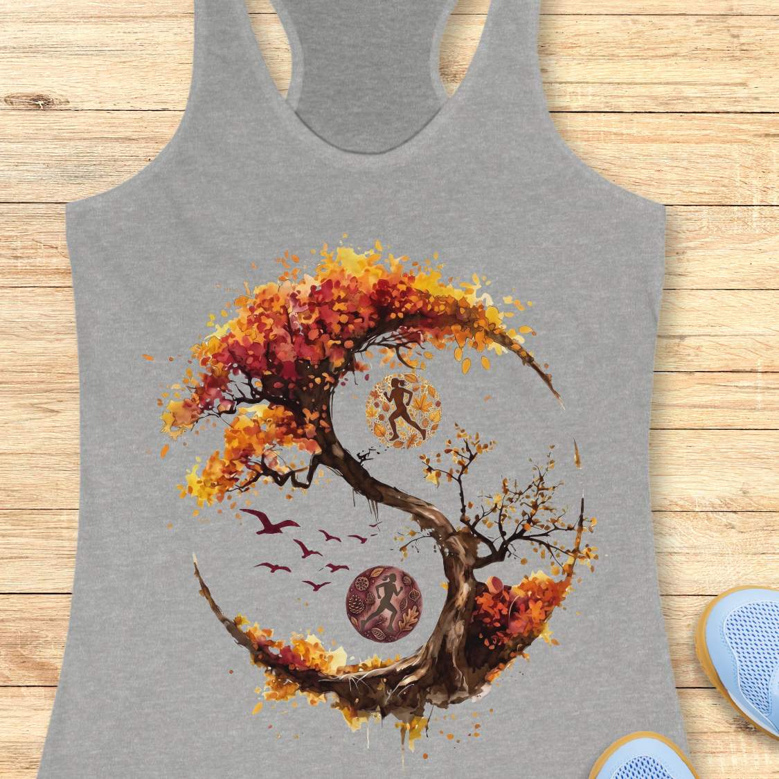 A heather gray racerback tank top featuring a large autumn-themed graphic print of a yin-yang tree with colorful fall leaves, birds, and a moon, displayed flat on a light wood background.