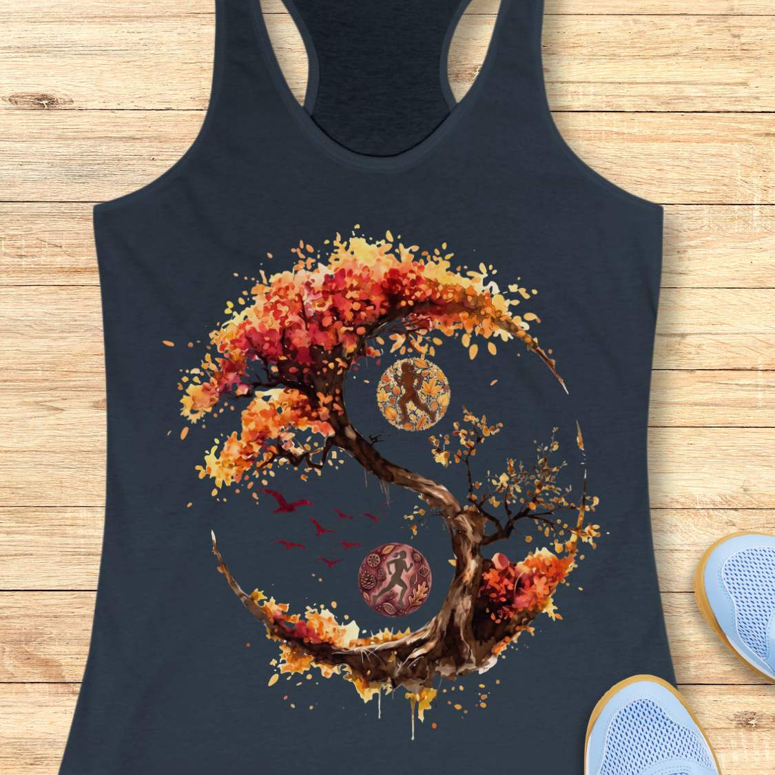 A indigo racerback tank top laid flat on a light wood plank background, featuring a large graphic of a stylized tree with autumn red, orange, and yellow leaves in a yin-yang shape. Part of a blue shoe is visible in the bottom right corner.