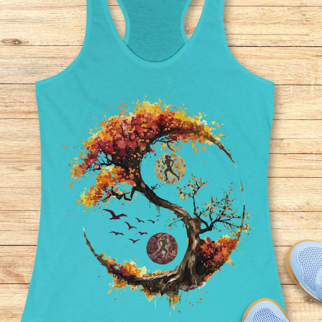 A blue racerback tank top laid flat on a light wood plank background, featuring a large graphic print of a stylized tree in autumn colors forming a circular yin-yang shape with a running figure, birds, and circular motifs. Part of a blue shoe is visible at the bottom right.