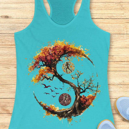 A blue racerback tank top laid flat on a light wood plank background, featuring a large graphic print of a stylized tree in autumn colors forming a circular yin-yang shape with a running figure, birds, and circular motifs. Part of a blue shoe is visible at the bottom right.