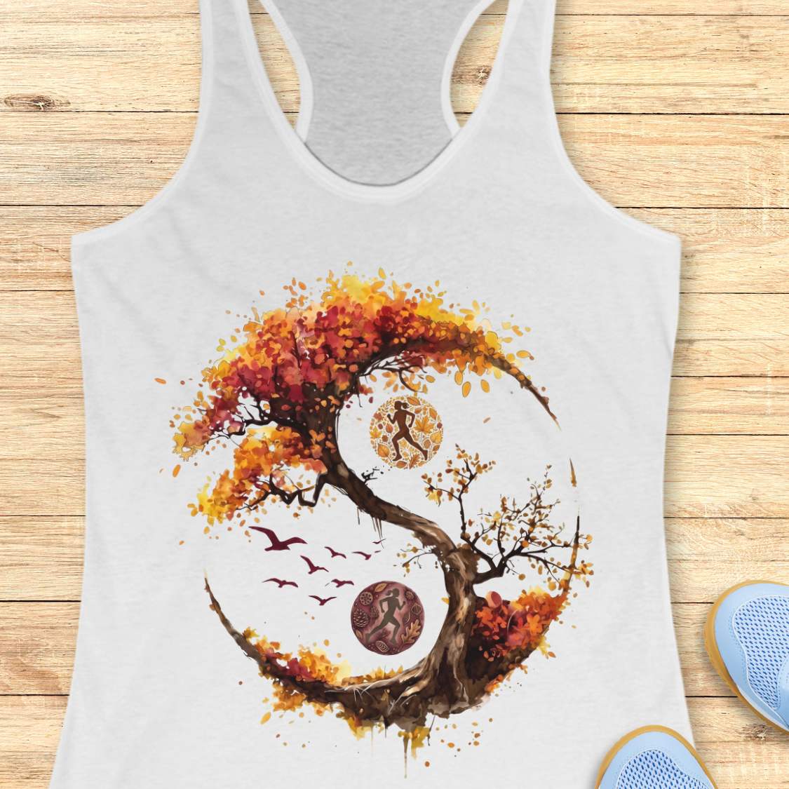 A white racerback tank top laid flat on a light wood plank background, featuring a large graphic print of a stylized tree in autumn colors forming a circular yin-yang shape with a running figure, birds, and circular motifs. Part of a blue shoe is visible at the bottom right.