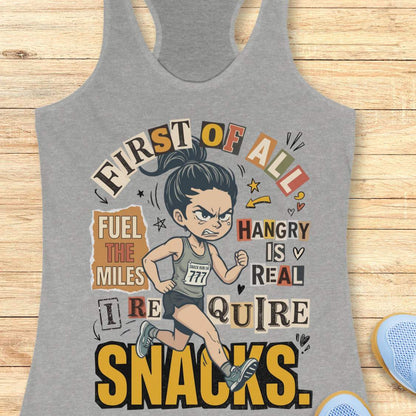 heather grey racerback tank top featuring a cartoon runner wearing a race bib and running shoes, surrounded by bold typography. The design reads: “First of all, fuel the miles. Hangry is real. I require snacks.” The artwork uses a playful mix of fonts, yellow, black, beige, and coral accents, with doodle-style arrows, stars, and hearts. The tank top is displayed on a light wood surface with running shoes partially visible in the corners.