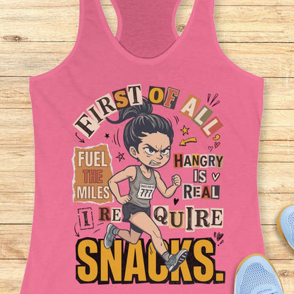 pink racerback tank top featuring a cartoon runner wearing a race bib and running shoes, surrounded by bold typography. The design reads: “First of all, fuel the miles. Hangry is real. I require snacks.” The artwork uses a playful mix of fonts, yellow, black, beige, and coral accents, with doodle-style arrows, stars, and hearts. The tank top is displayed on a light wood surface with running shoes partially visible in the corners.
